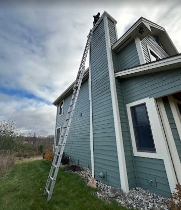 Tall ladder against a two-story house with a person working near the roof, cloudy sky in the background.