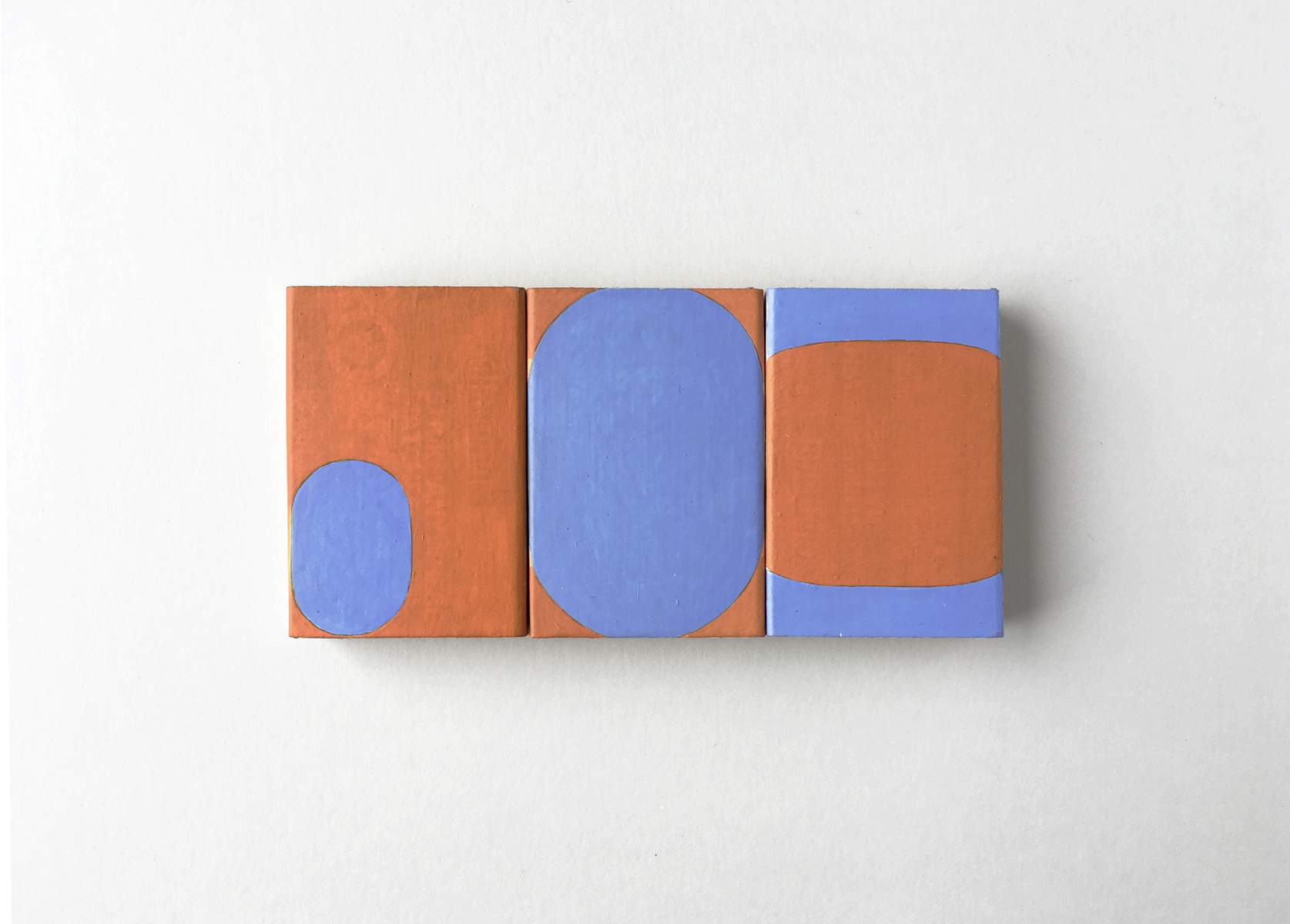 Three small matchboxes in a row with blue and orange ovals painted on them.
