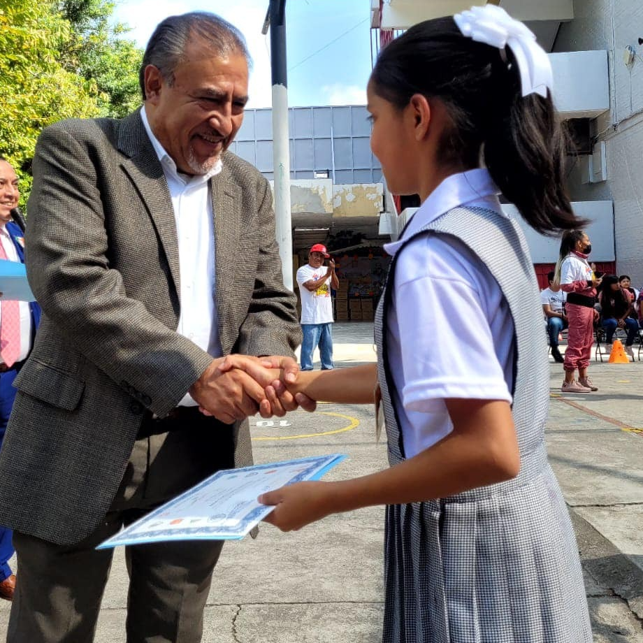 Hombre de negocios sonriendo, vestido con traje gris y corbata azul, sentado en una oficina moderna iluminada por una lámpara.