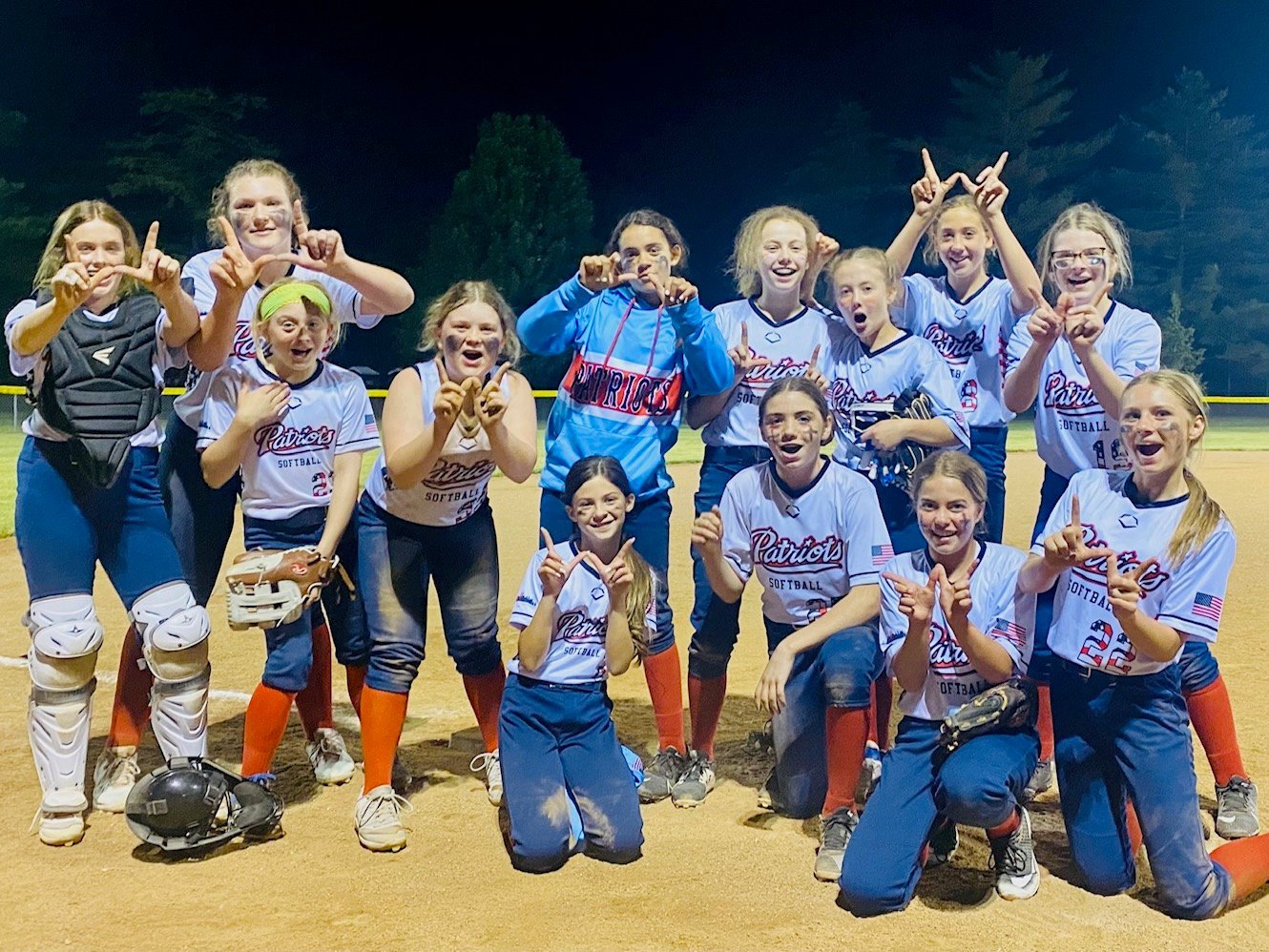 Youth softball team celebrating a win at night on the field, wearing Patriots uniforms with protective gear.