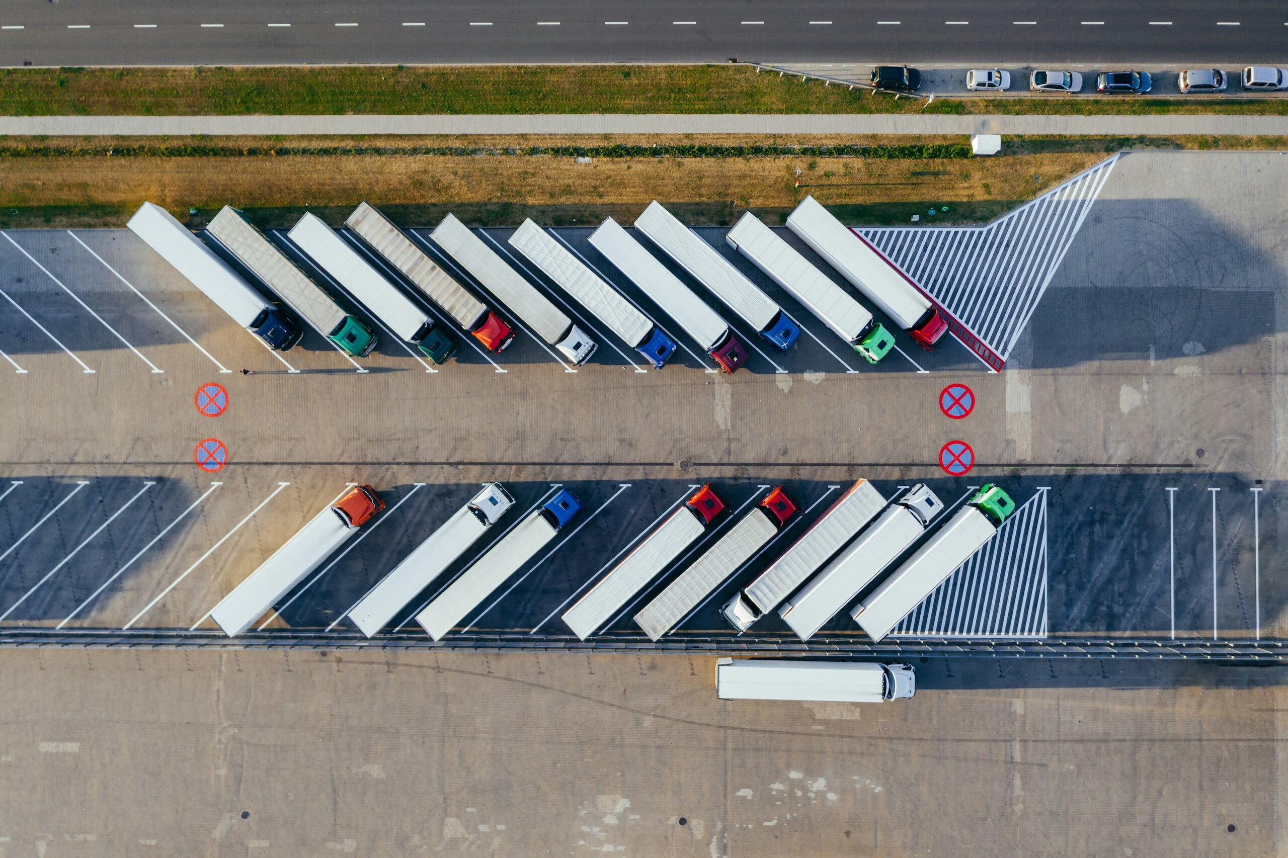 Vista aérea de un estacionamiento de trailers con varios camiones alineados diagonalmente, áreas marcadas y coches estacionados al fondo.