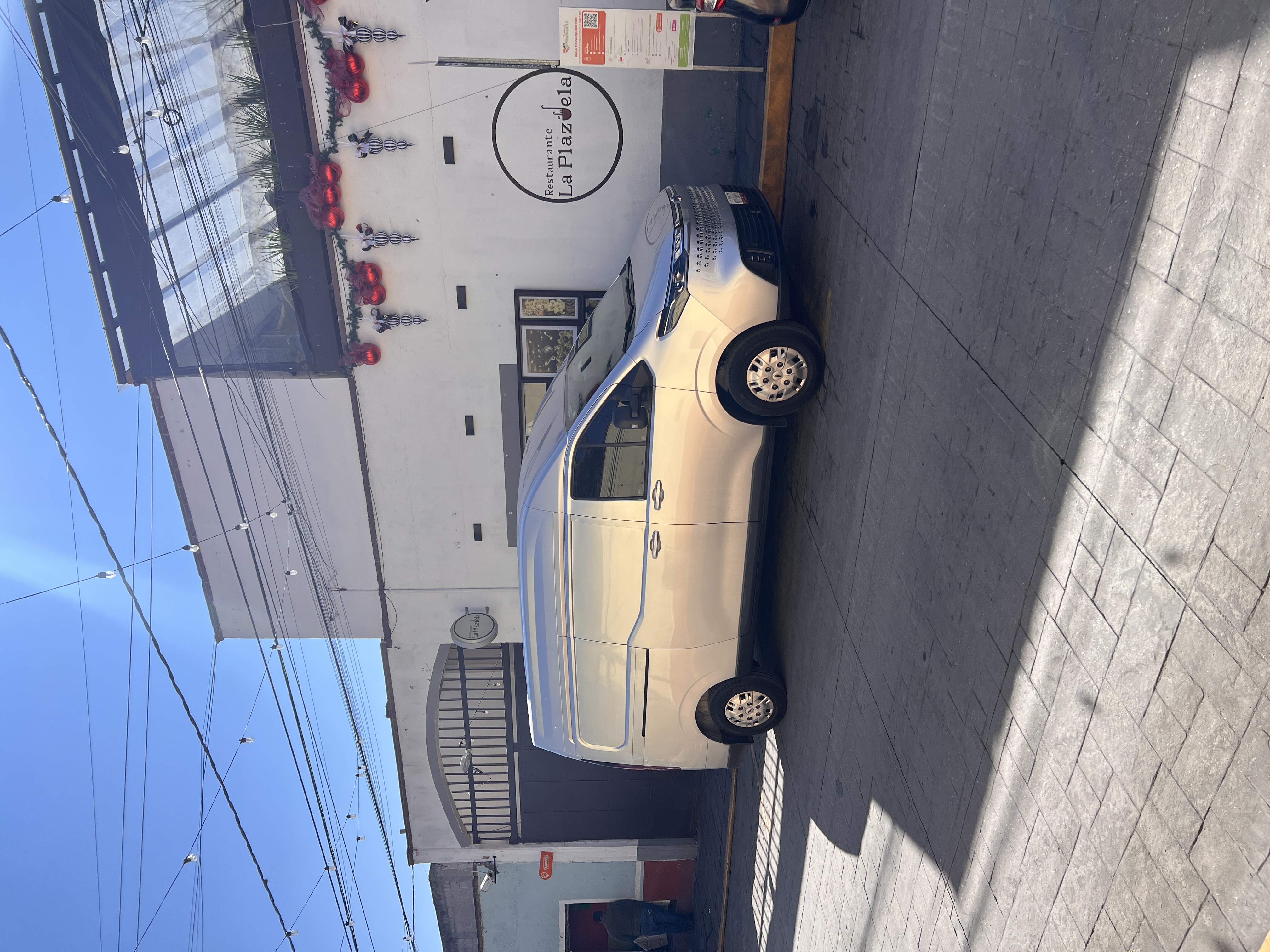 Silver van parked in front of a restaurant with a sign reading 'Restaurante La Plazuela' and decorative outdoor string lights.