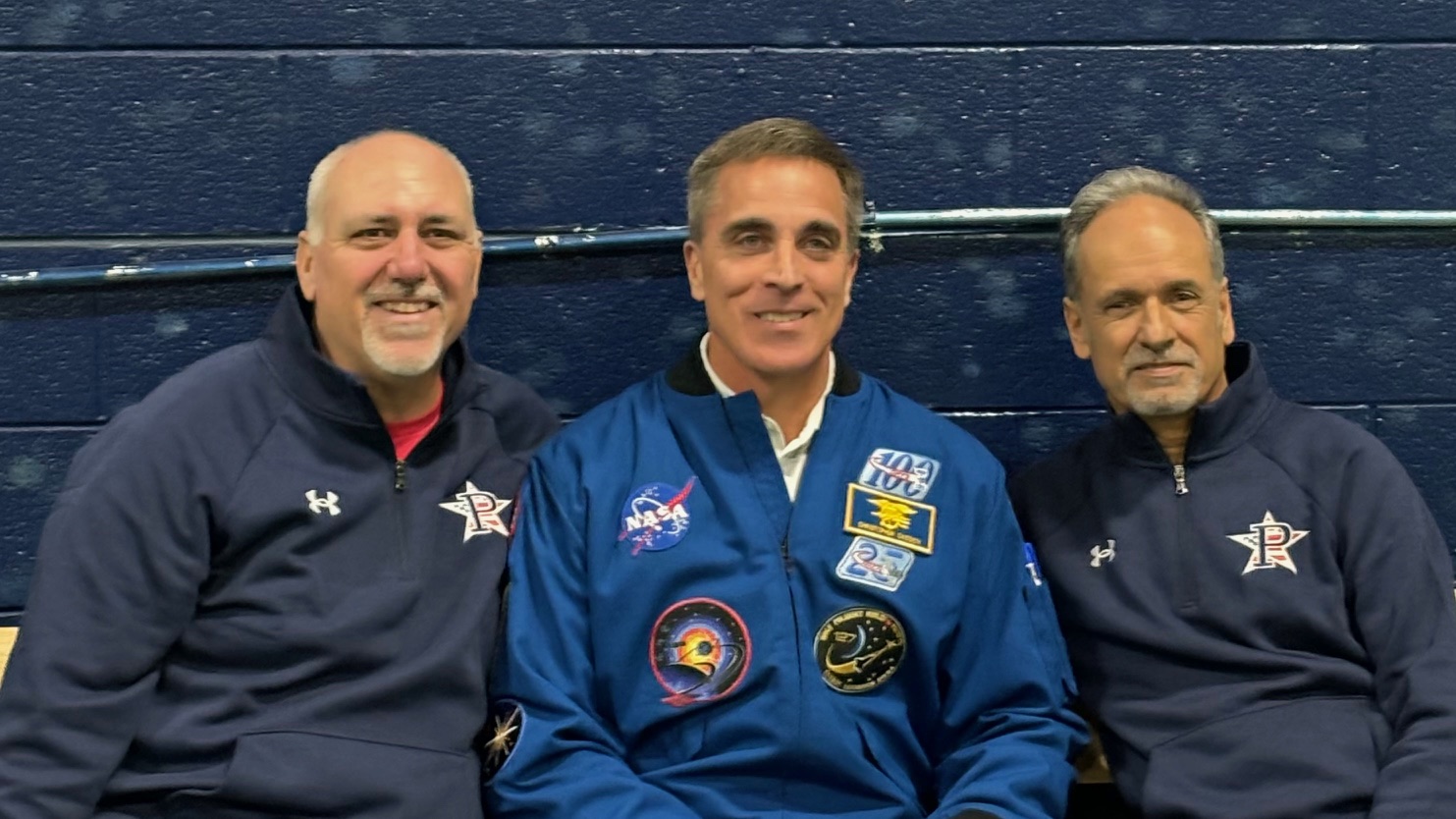 Three men smiling, center man in blue NASA jacket with mission patches, seated against a dark blue wall.