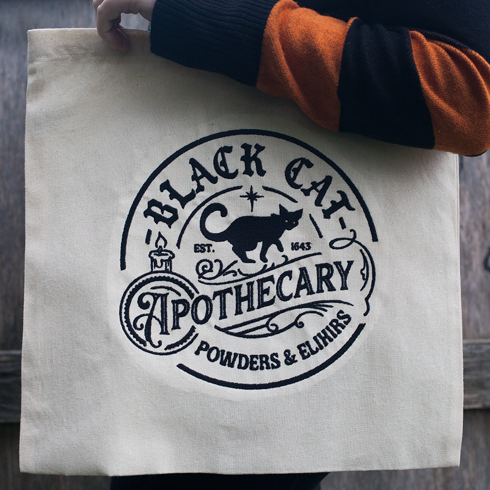 A white tote bag featuring an embroidered black cat apothecary design