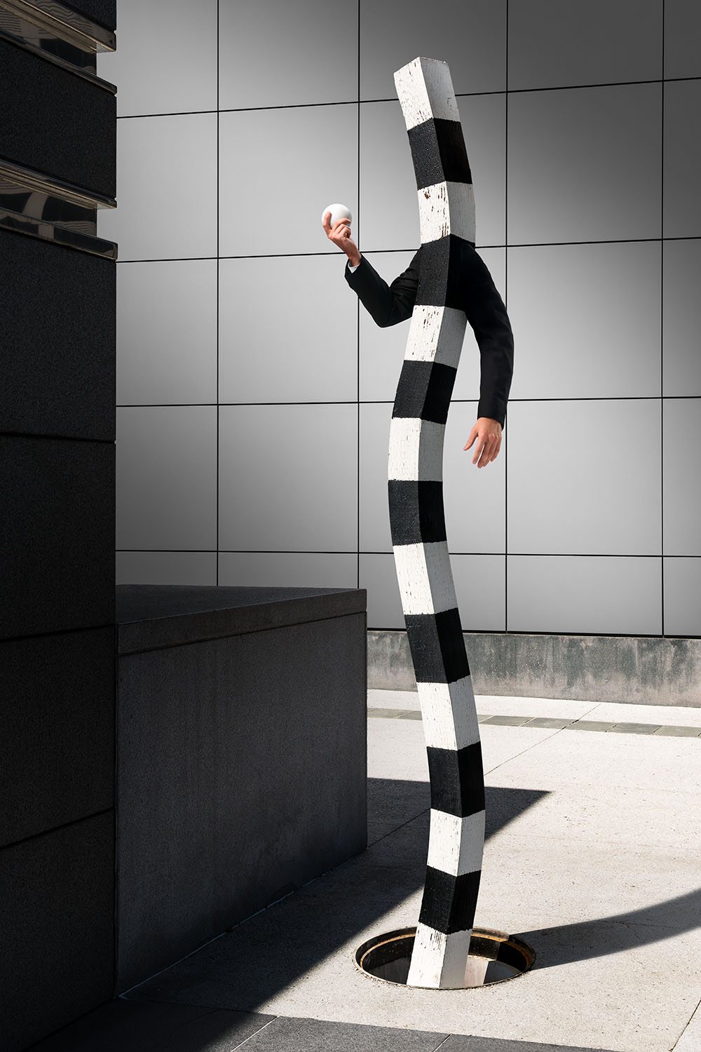 Fantastical creature with a body of a black and white striped wooden post and the arms of a man, emerging from a manhole. 