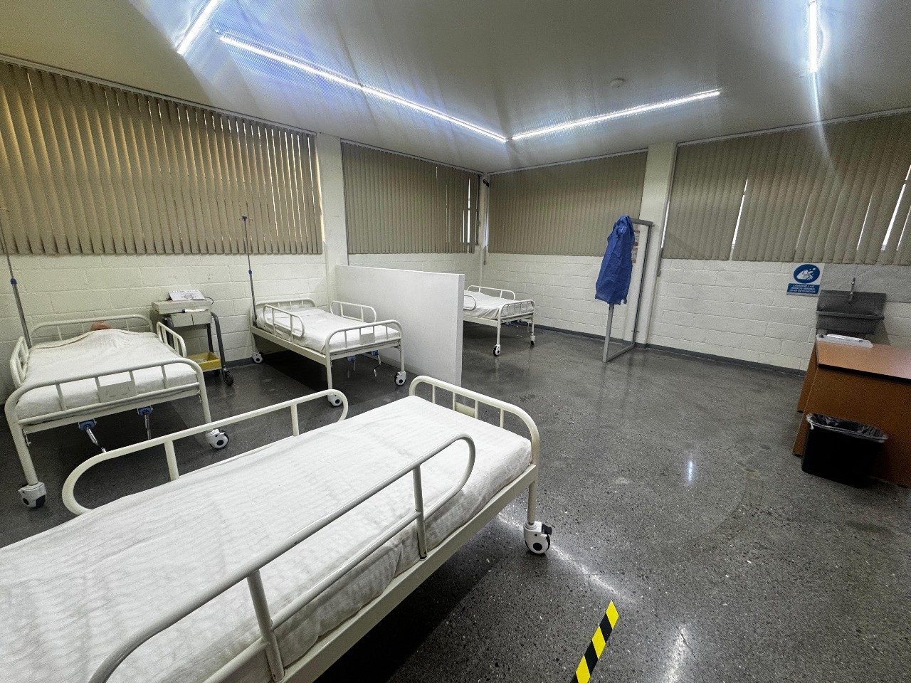 Sala de hospital vacía con camas blancas, cortinas cerradas y luz fluorescente.
