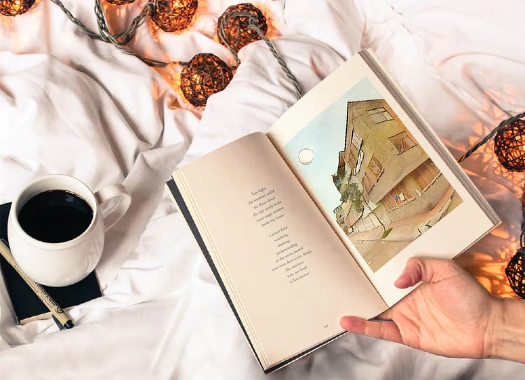 Mano sosteniendo un libro abierto con poema y dibujo, taza de café y luces decorativas sobre una manta blanca.