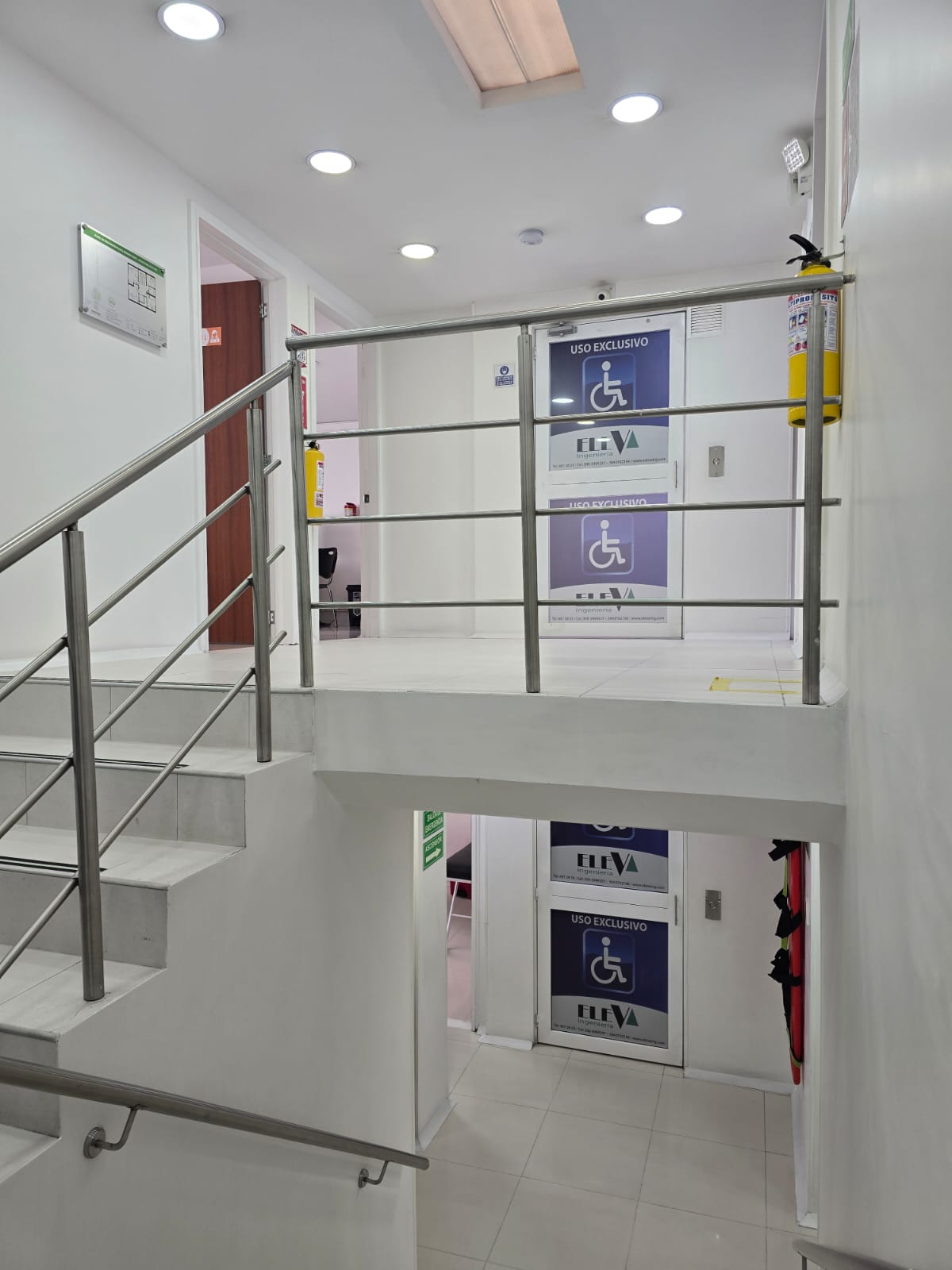Interior de un edificio moderno con escaleras y puertas de elevador marcadas para uso exclusivo de personas con discapacidad, señaladas con carteles azules.