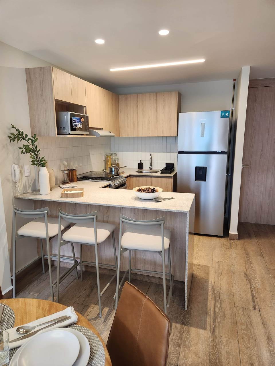 Cocina moderna con muebles de madera clara, isla con taburetes, electrodomésticos de acero inoxidable y decoración minimalista.