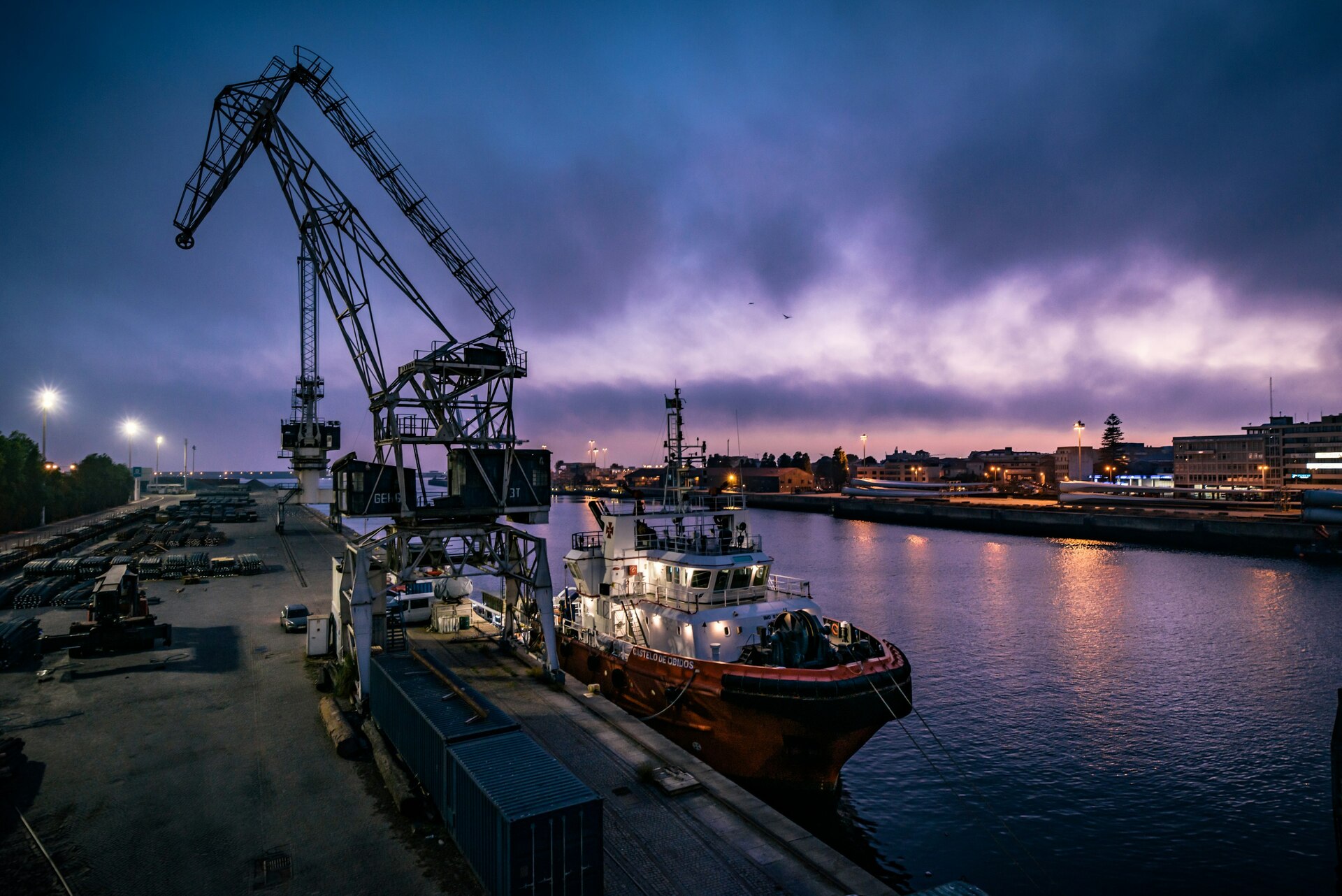 Grúa portuaria y barco atracado en el puerto al atardecer con luces urbanas reflejándose en el agua. Grúa portuaria y barco atracado en el puerto al atardecer con luces urbanas reflejándose en el agua.