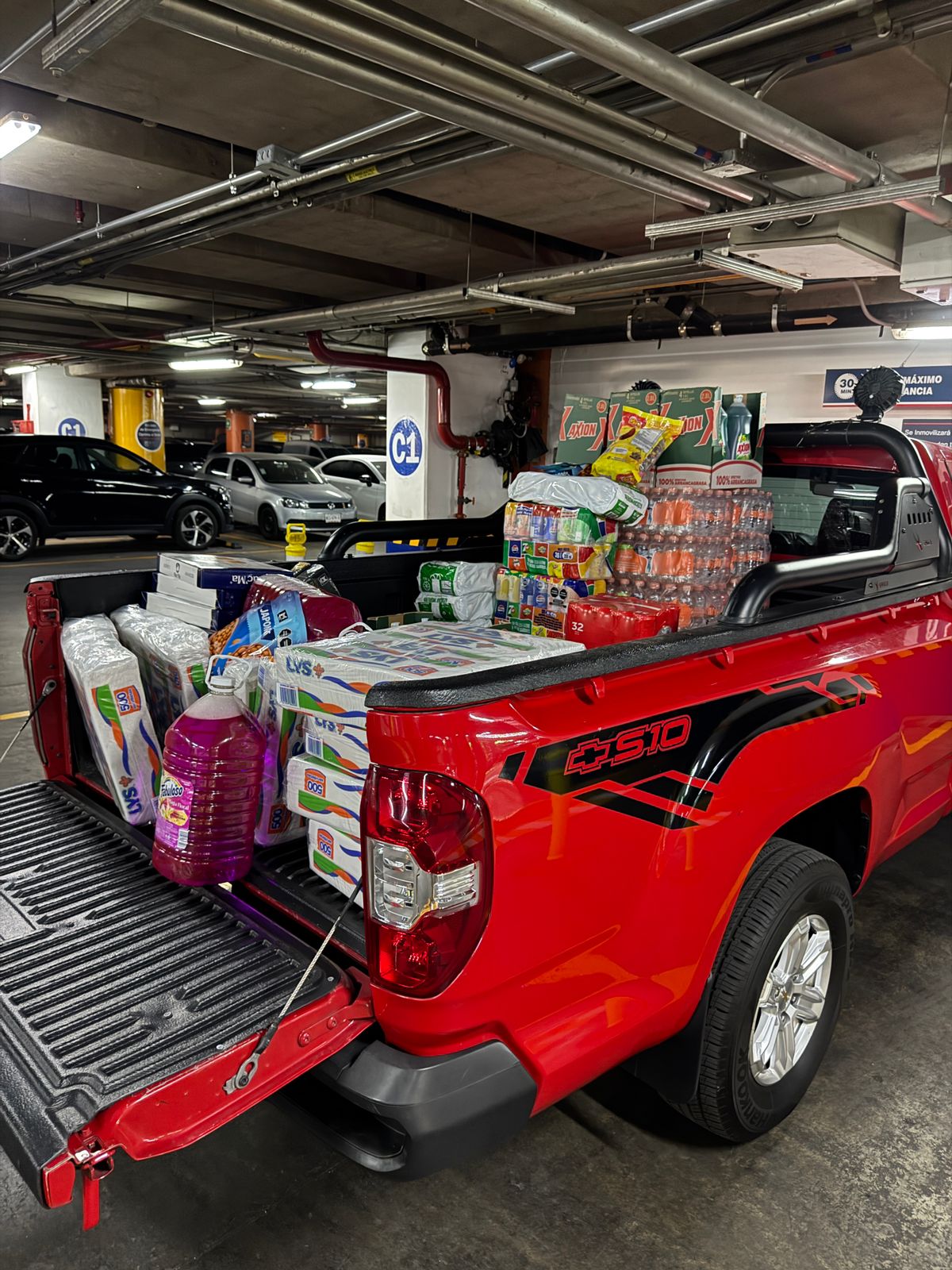 Red pickup truck loaded with bulk household supplies, including paper towels, drinks, and cleaning products, parked in an underground garage.
