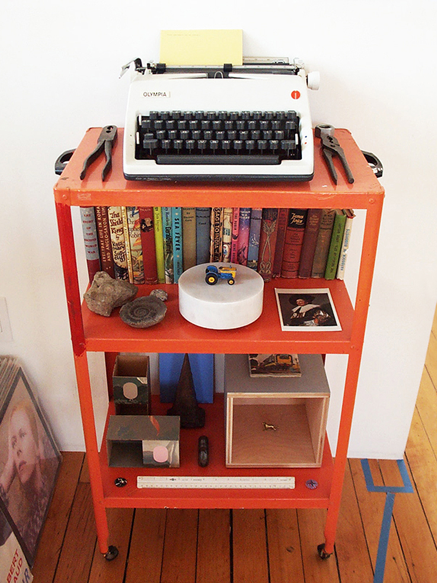A typewriter, books, Matchbox tractor and other objects on an orange utility cart.