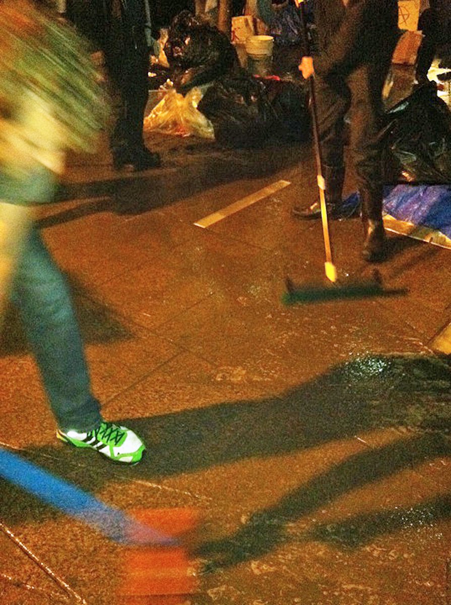 Two men scrub at a wet paved area with brooms, at night in orange streetlight.