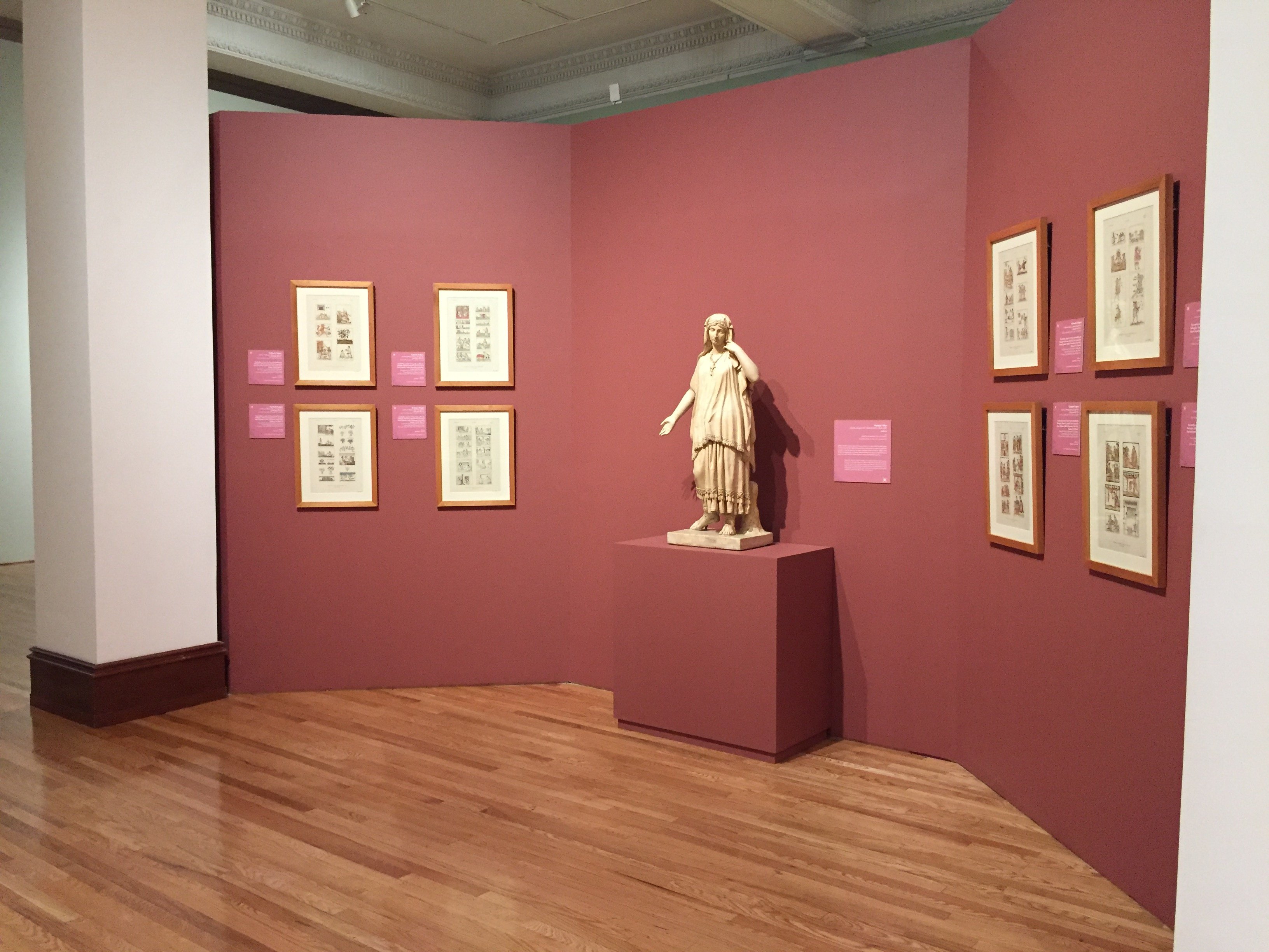 Exposición de arte en museo con una estatua central y varias ilustraciones enmarcadas sobre paredes color rosa.