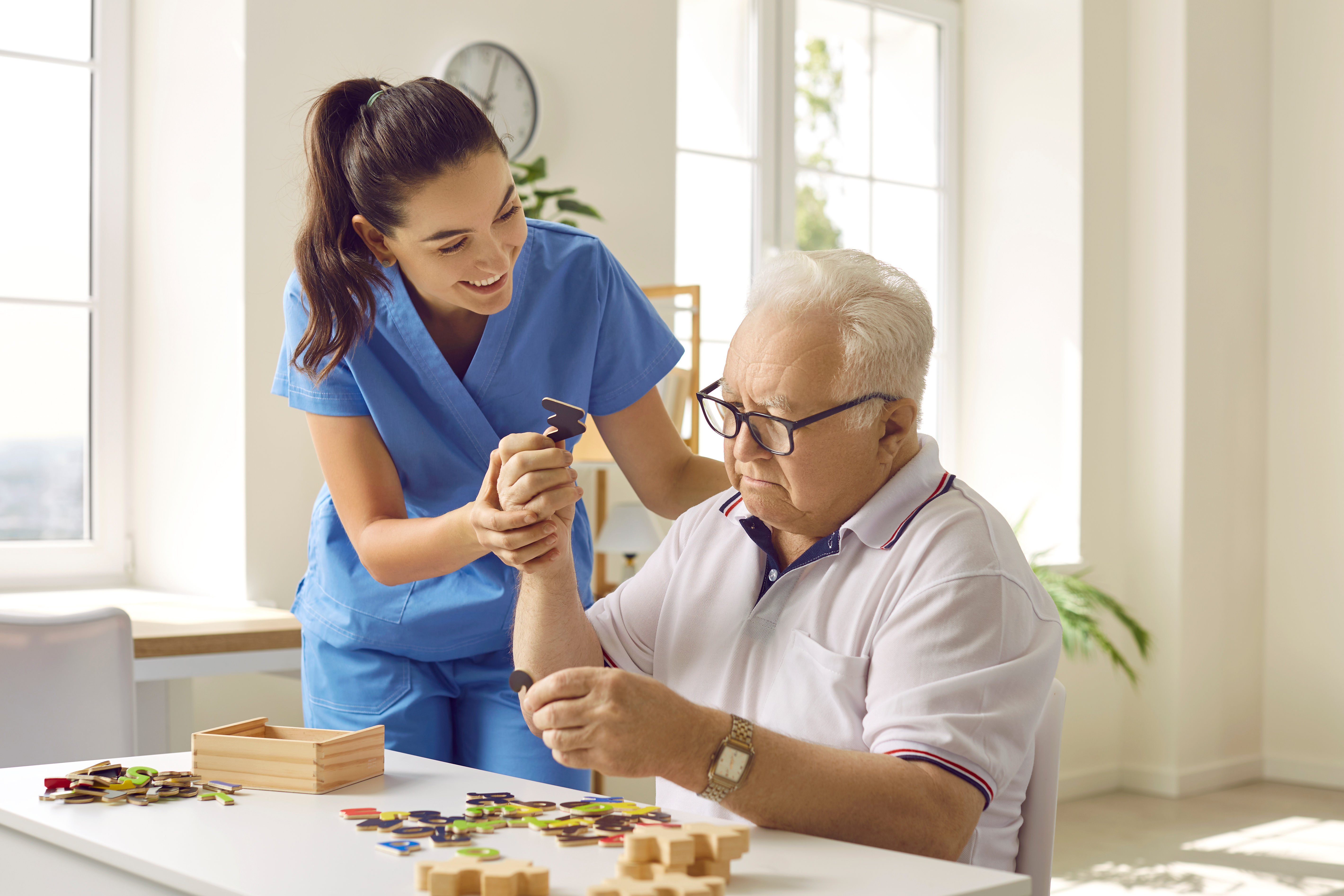 Female caregiver in blue scrubs assists elderly man with puzzle pieces at a table in a bright, sunny room.