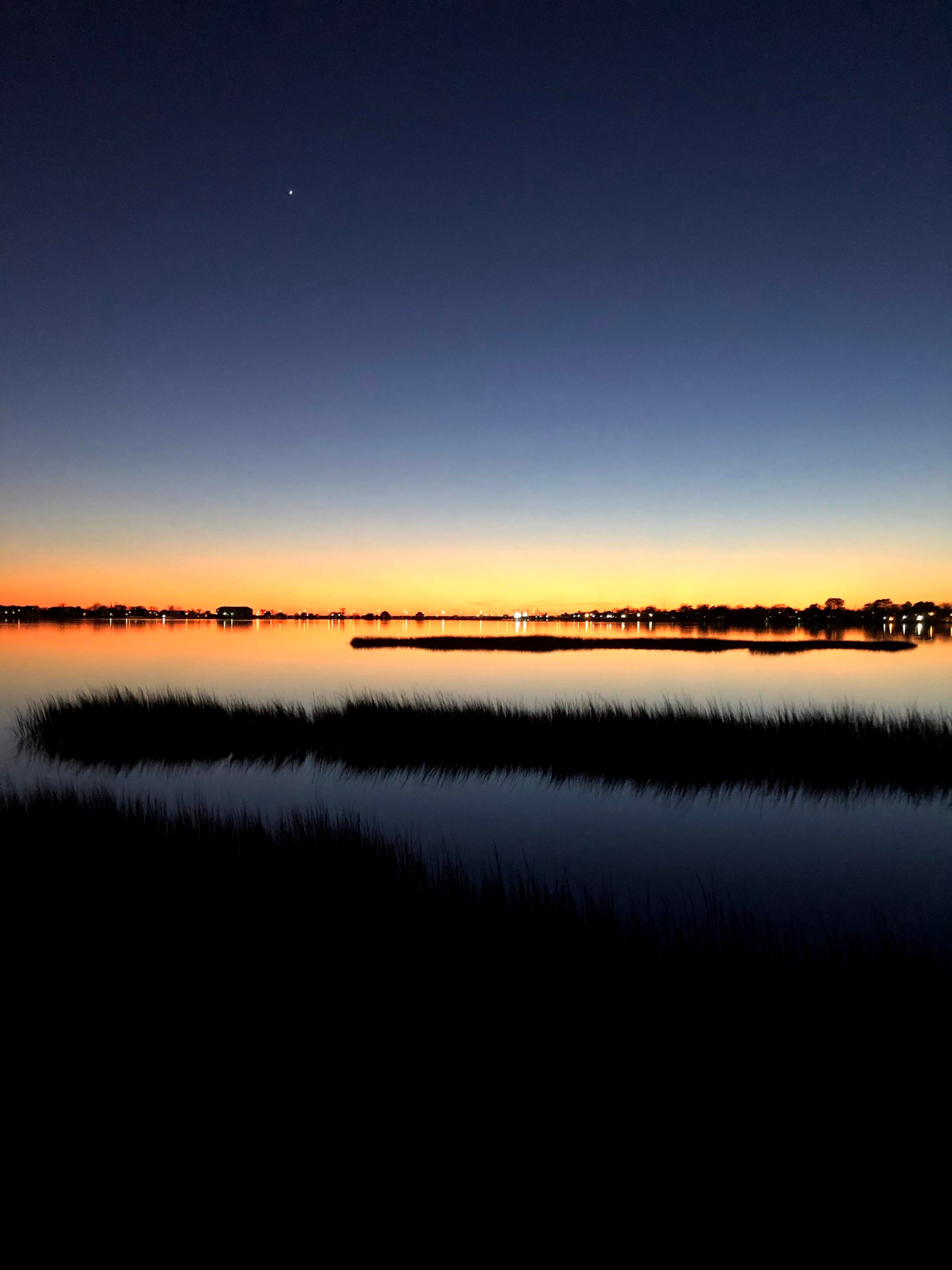 Tranquil sunset over a calm lake with silhouetted reeds and clear sky.
