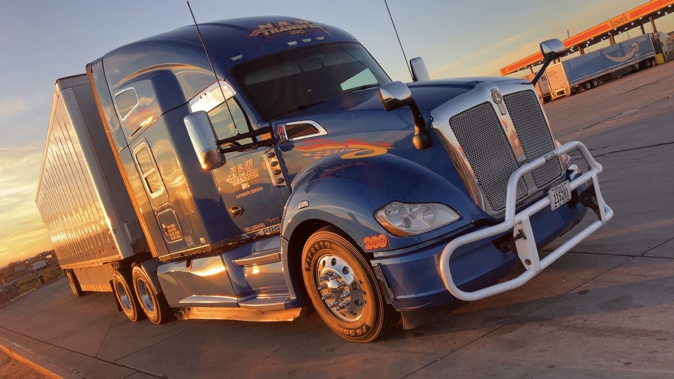 Blue semi-truck parked at sunset with orange hues on the horizon, featuring chrome details and N & W Transfer Inc. branding.
