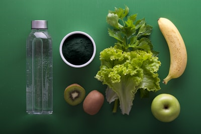 Assorted healthy foods on a green background, including a water bottle, spirulina powder, lettuce, celery leaves, banana, kiwi, potato, and green apple. Assorted healthy foods on a green background, including a water bottle, spirulina powder, lettuce, celery leaves, banana, kiwi, potato, and green apple.