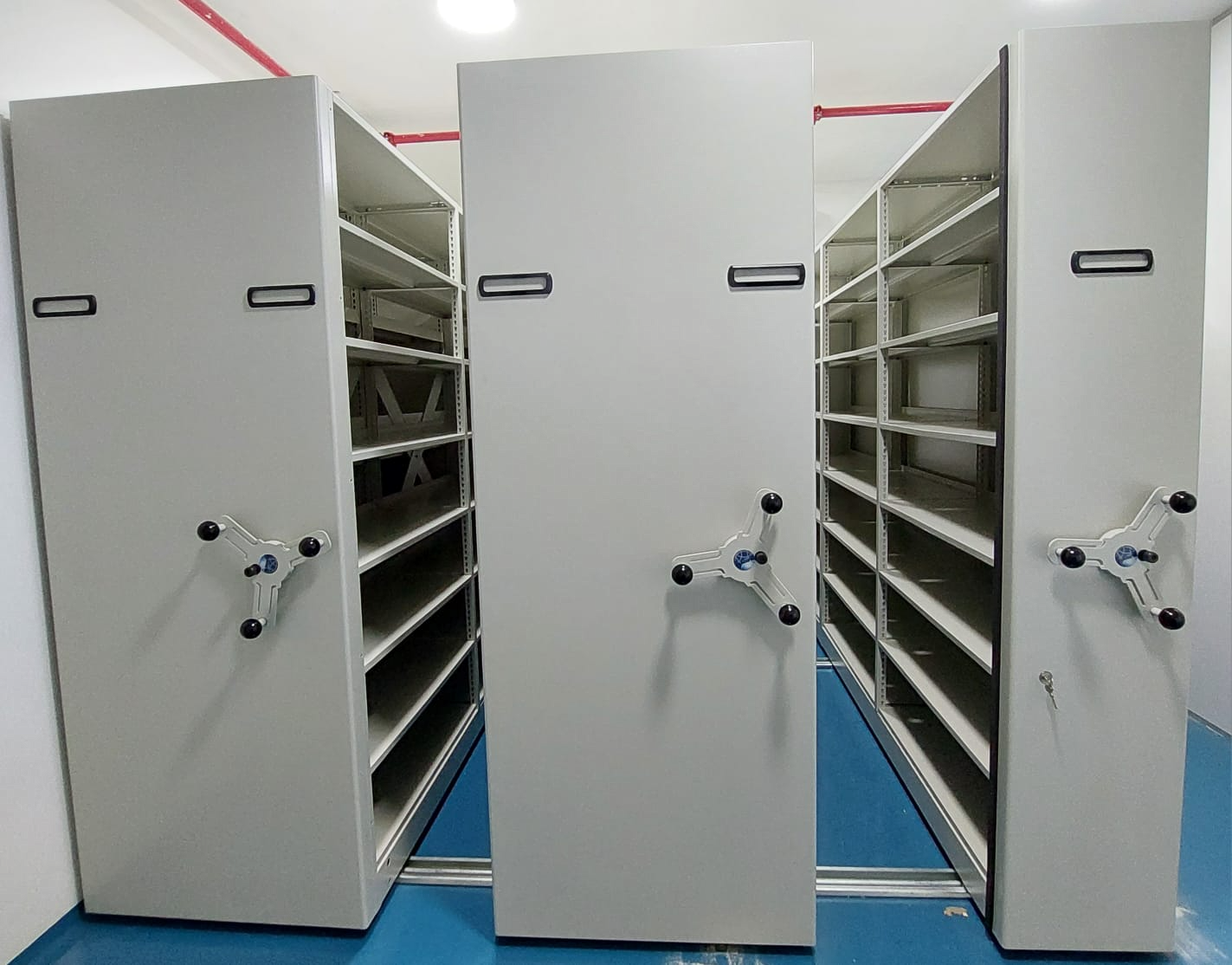 Estante deslizante de arquivo cinza em ambiente corporativo, com pisos azuis e alças para movimentação.