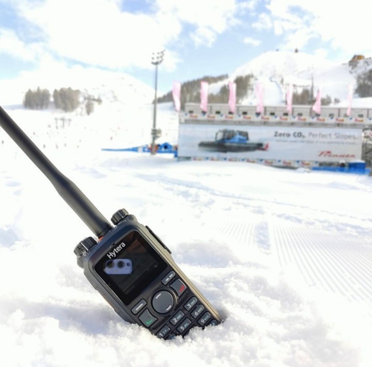 A Hytera radio partially buried in snow at a ski resort with snowy mountains and a billboard in the background.