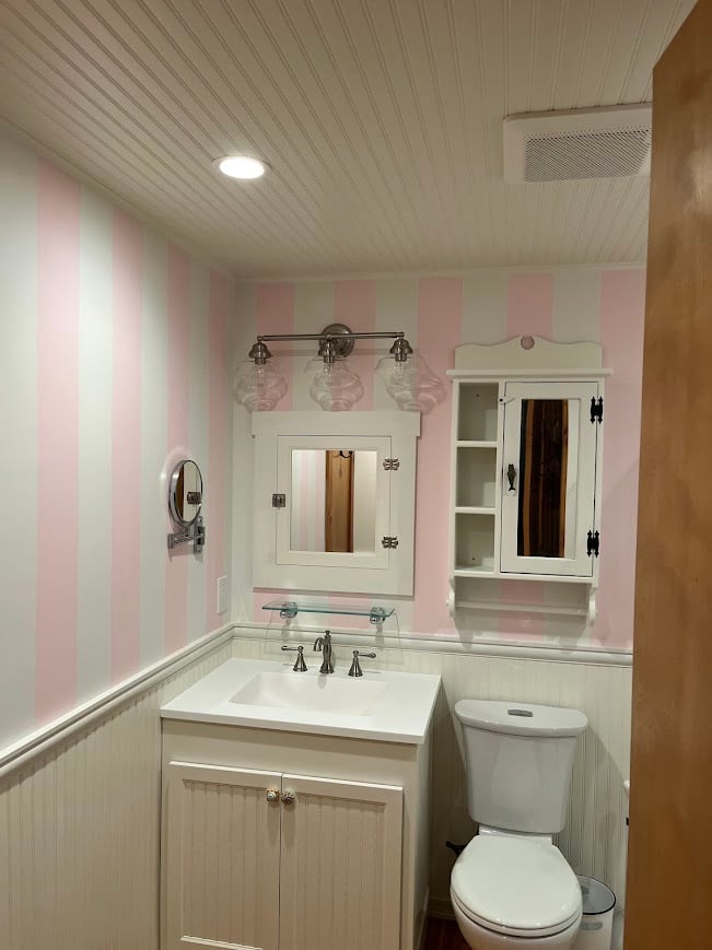 Small bathroom with pink and white striped walls, white vanity with sink, mirrored cabinet, toilet, and decorative lighting.
