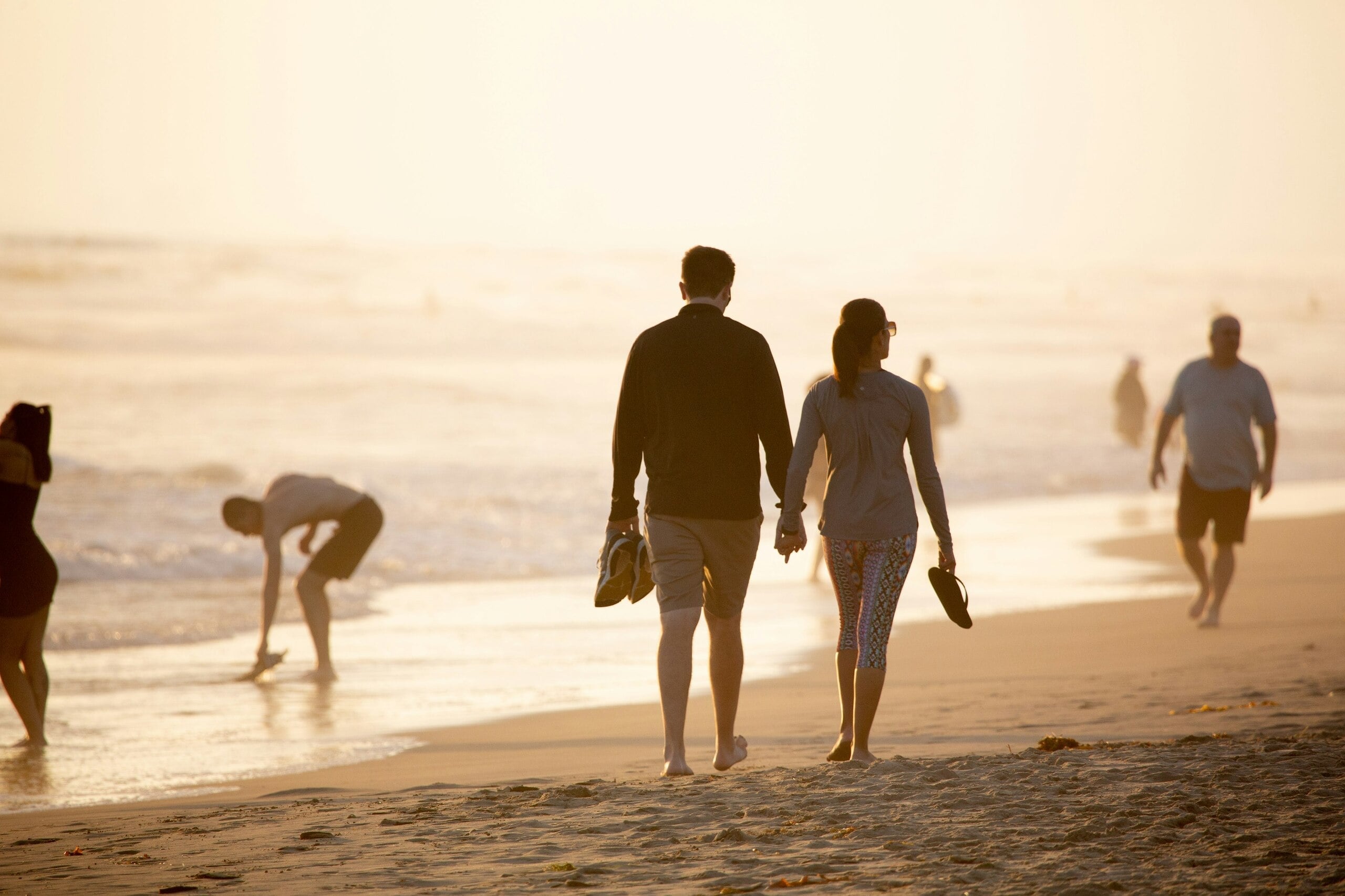 Pareja caminando de la mano por la playa al atardecer con personas junto al mar