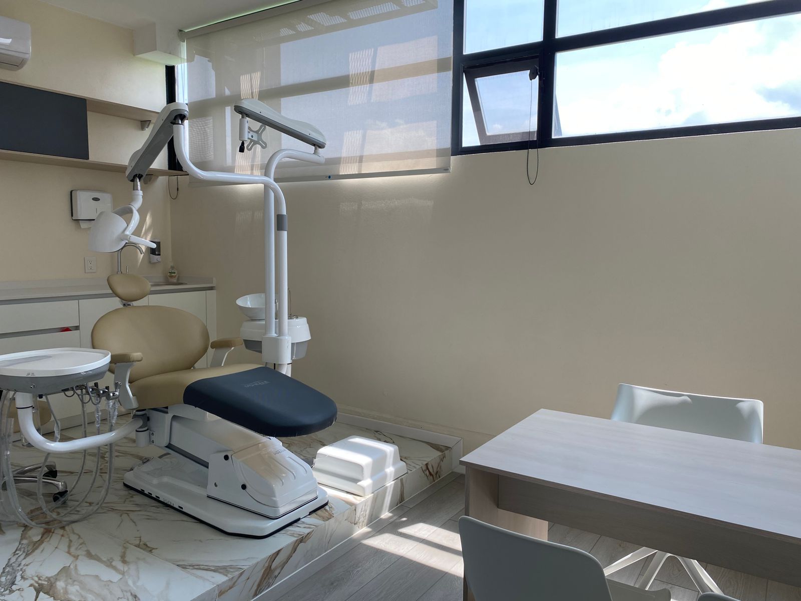 Consultorio dental moderno con silla odontológica beige, lámpara de examen y silla de escritorio junto a gran ventana.