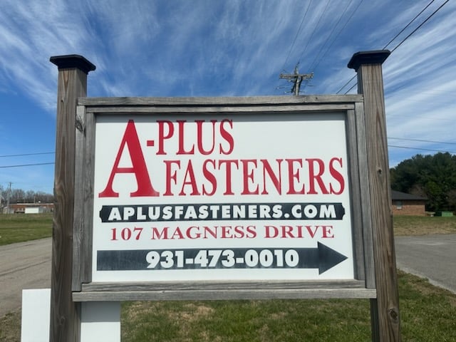 Sign for A-Plus Fasteners with website, address, and phone number on a wooden frame against a blue sky.
