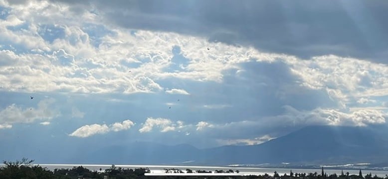 Paisaje natural con cielo nublado y rayos de sol filtrándose, reflejando sobre un lago sereno, con montañas en el fondo.