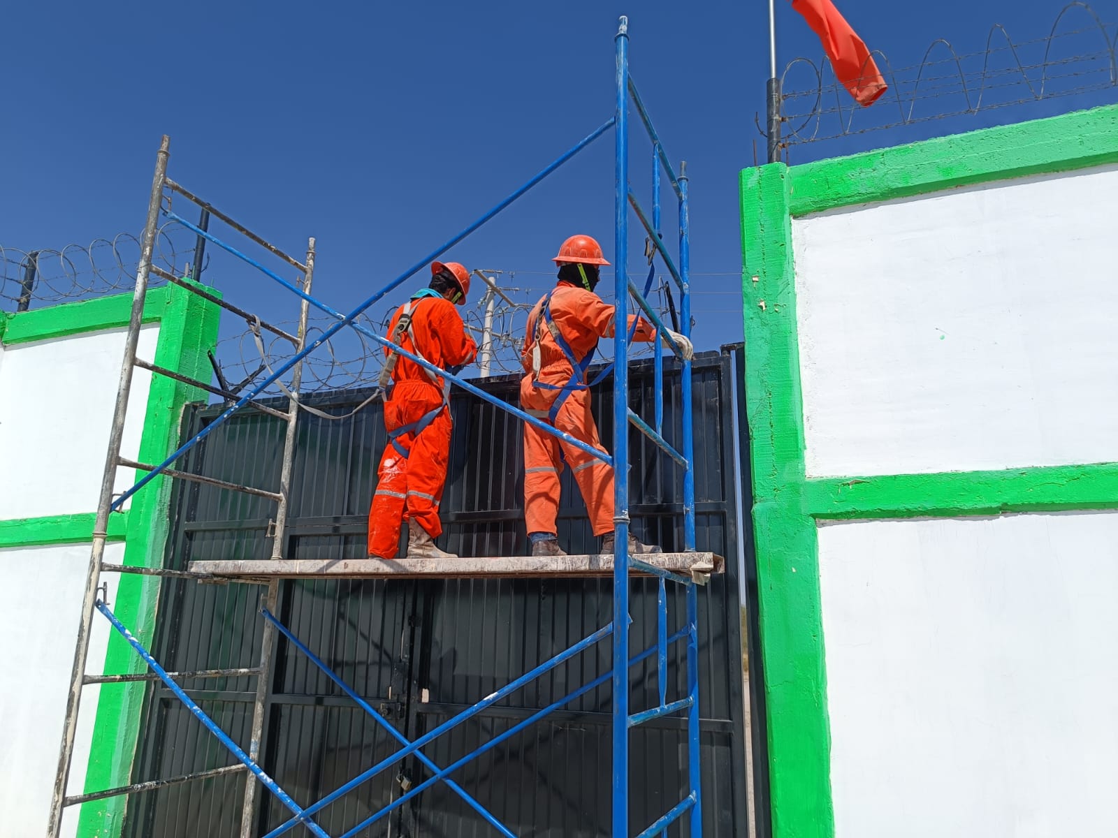 Trabajadores de construcción en andamios, vistiendo trajes naranjas y cascos, pintando una pared blanca con borde verde, con cielo azul de fondo.