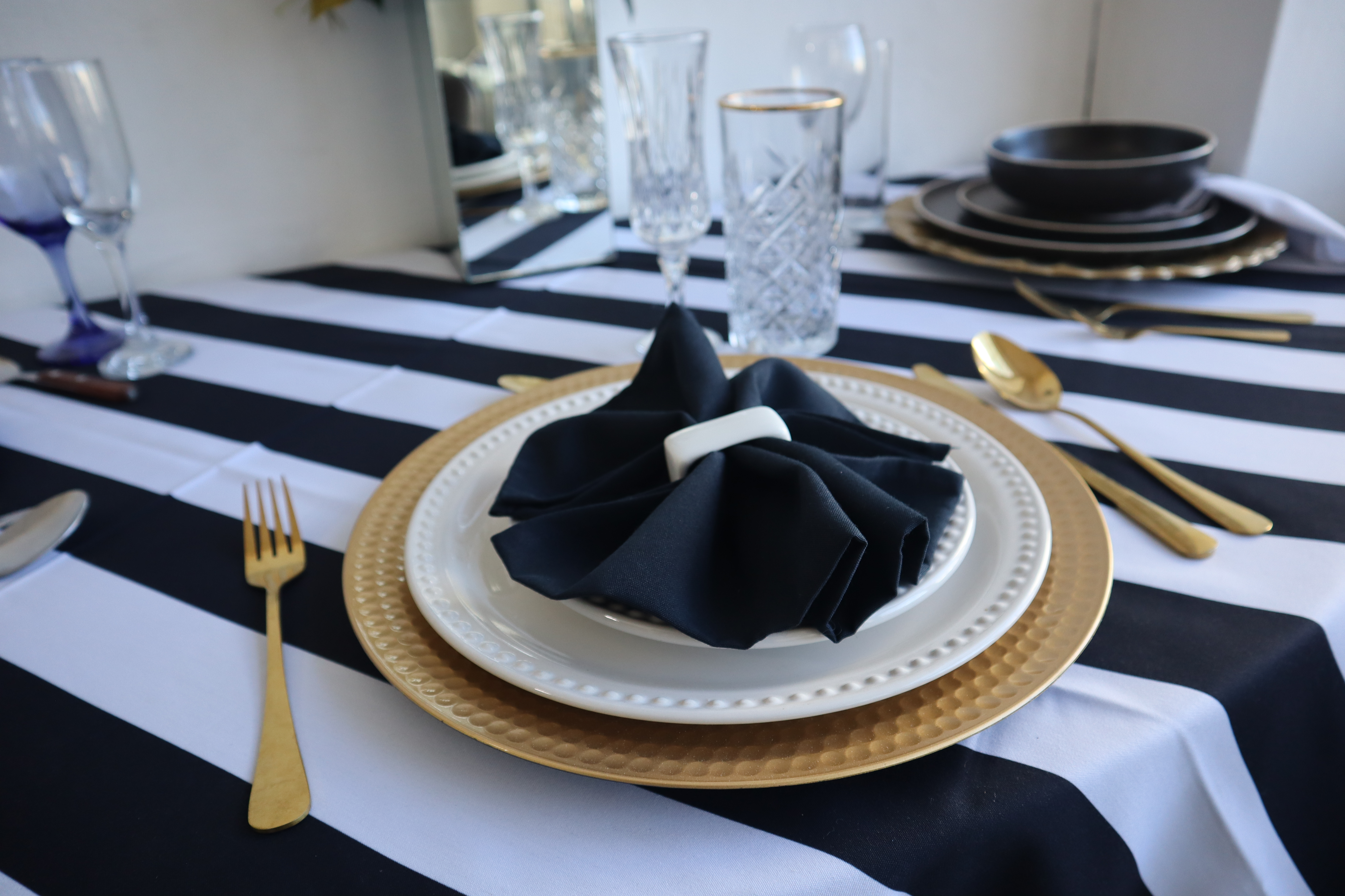 Mesa elegante con vajilla blanca y dorada, servilleta negra, utensilios dorados y mantel a rayas blanco y negro. Mesa elegante con vajilla blanca y dorada, servilleta negra, utensilios dorados y mantel a rayas blanco y negro.