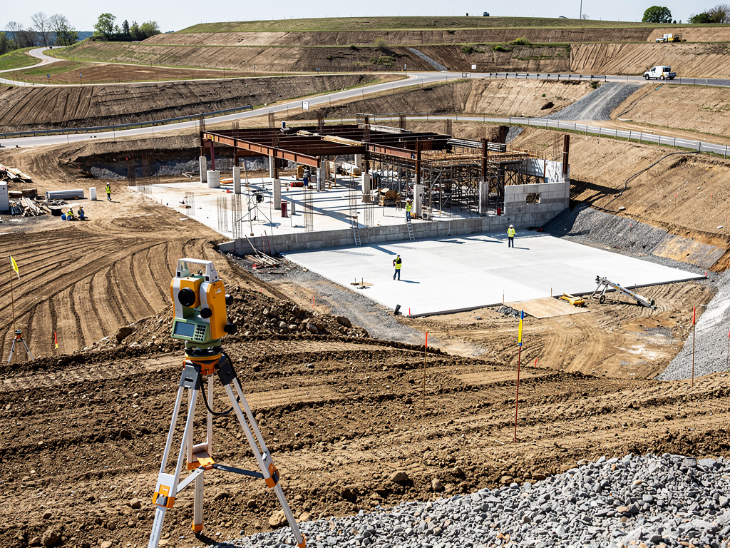 Obra de construcción con estructura de acero, losa de hormigón y estación total topográfica en primer plano