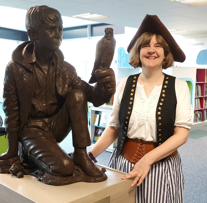 Author Susan Brownrigg at Hoyland Library. She is wearing a pirate costume and is stood next to the sculpture of Billy Casper and Kes, the kestrel, from the book A Kestrel for a Knave.