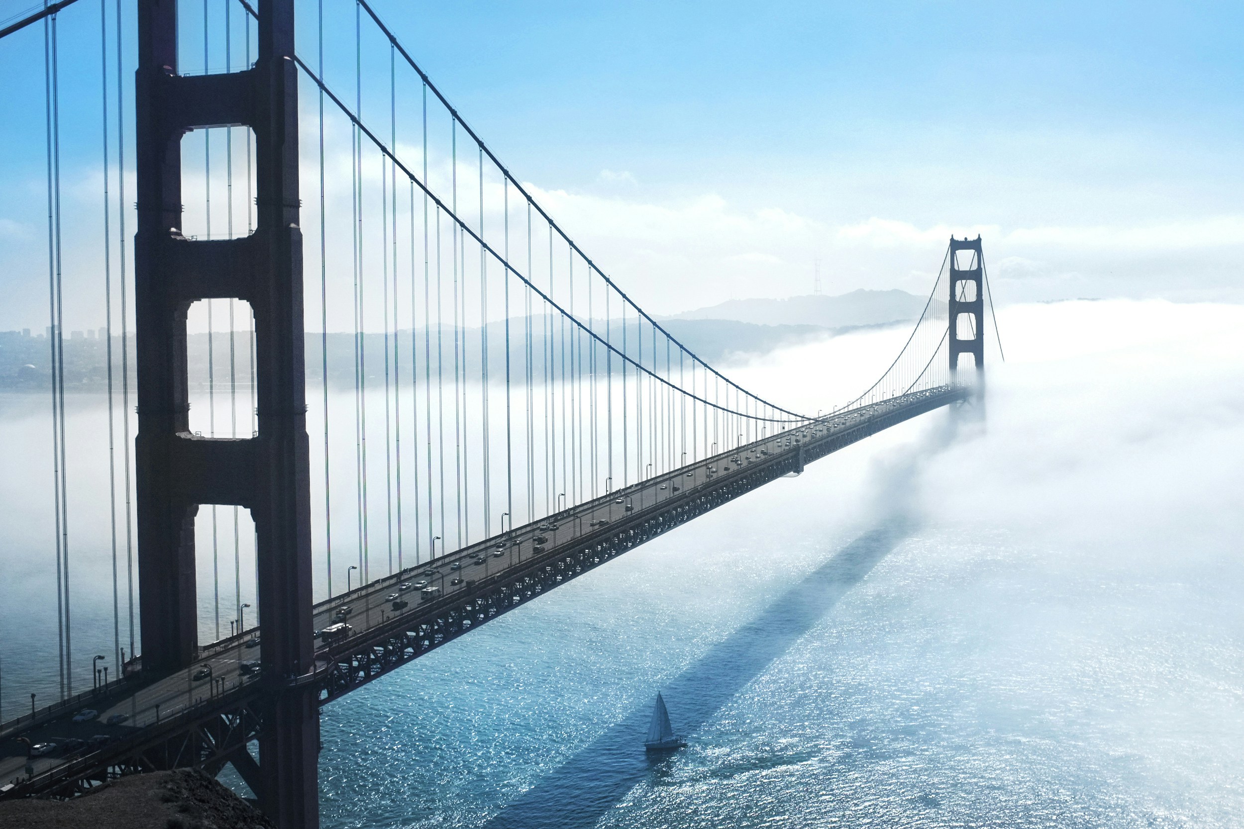 Golden Gate Bridge shrouded in fog with a sailboat on the water below and clear blue sky above.